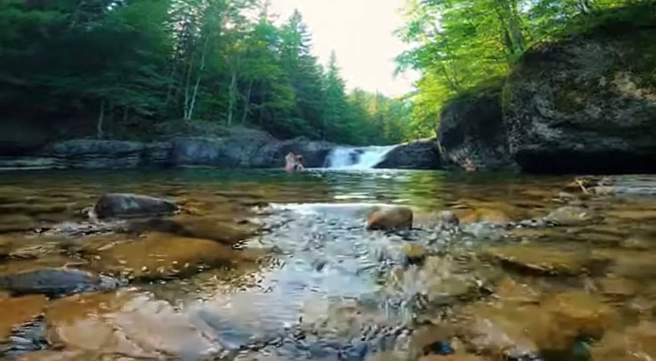 Clear New England swimming hole with amber river stones, a low waterfall, and forested rock walls in summer light.