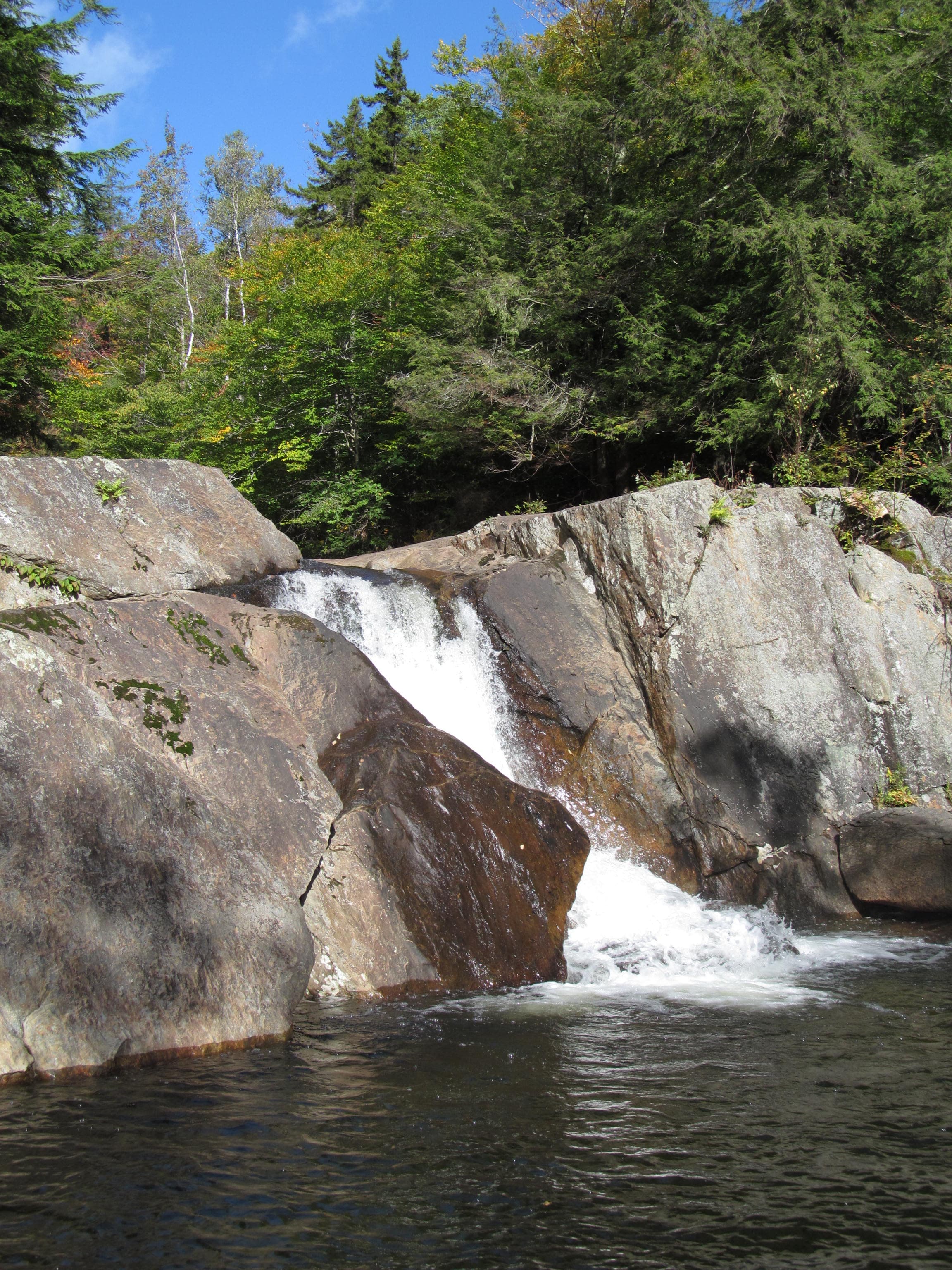 Cascade between granite boulders into a dark pool, green forest and blue sky, Buttermilk Falls, Ludlow, Vermont.