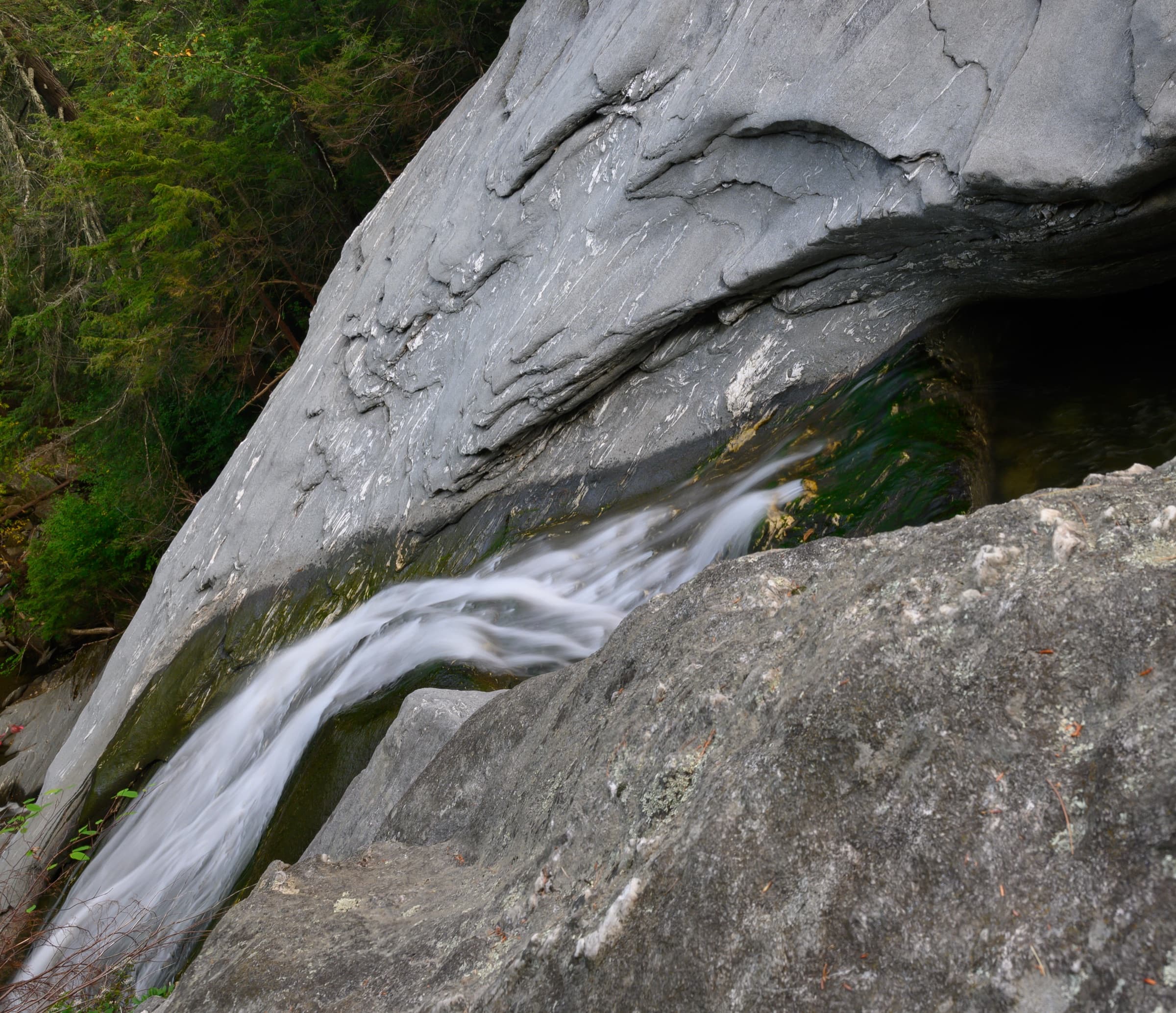 Water slipping through a narrow rock chute beneath a smooth gray slab at Hamilton Falls.